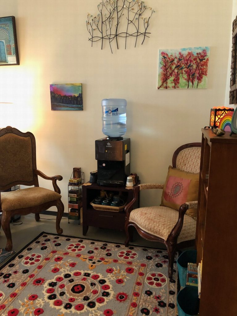 Two upholstered armchairs on a red and gray patterned rug, beneath colorful paintings